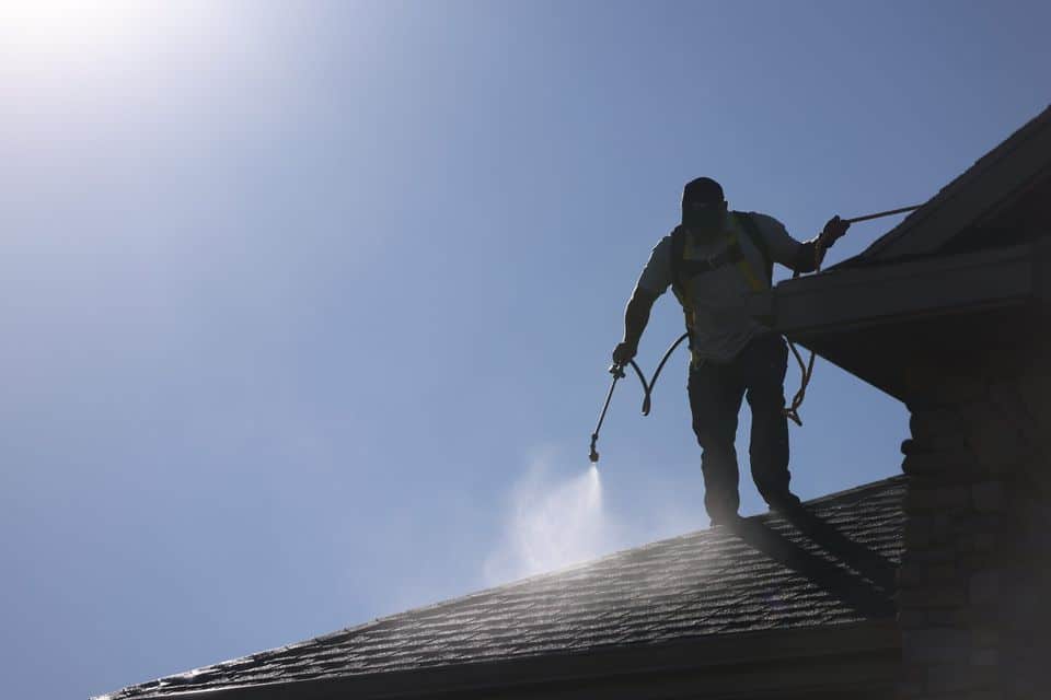 Technician in a safety harness applying restoration treatment from a hose along the eaves of an asphalt shingle roof.