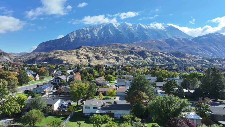 Drone view over a fall-color neighborhood toward snowcapped Wasatch peaks. The NuRoof Guys.