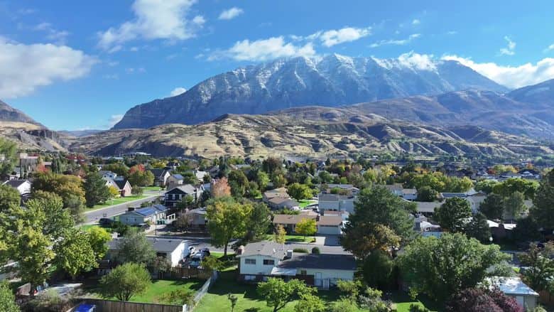Drone gliding over a fall-color neighborhood toward snowcapped Wasatch peaks. The NuRoof Guys.