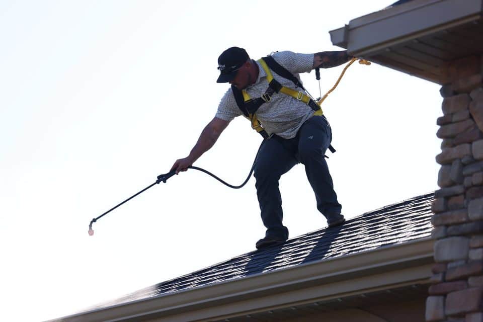 Technician in a yellow safety harness with a tie-off line, applying GoNano roof restoration treatment from a hose along an asphalt shingle roof edge.