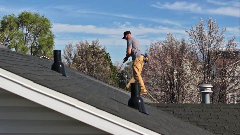 Technician applying treatment from a hose line on a gray shingle roof, with flowering trees behind. The NuRoof Guys.